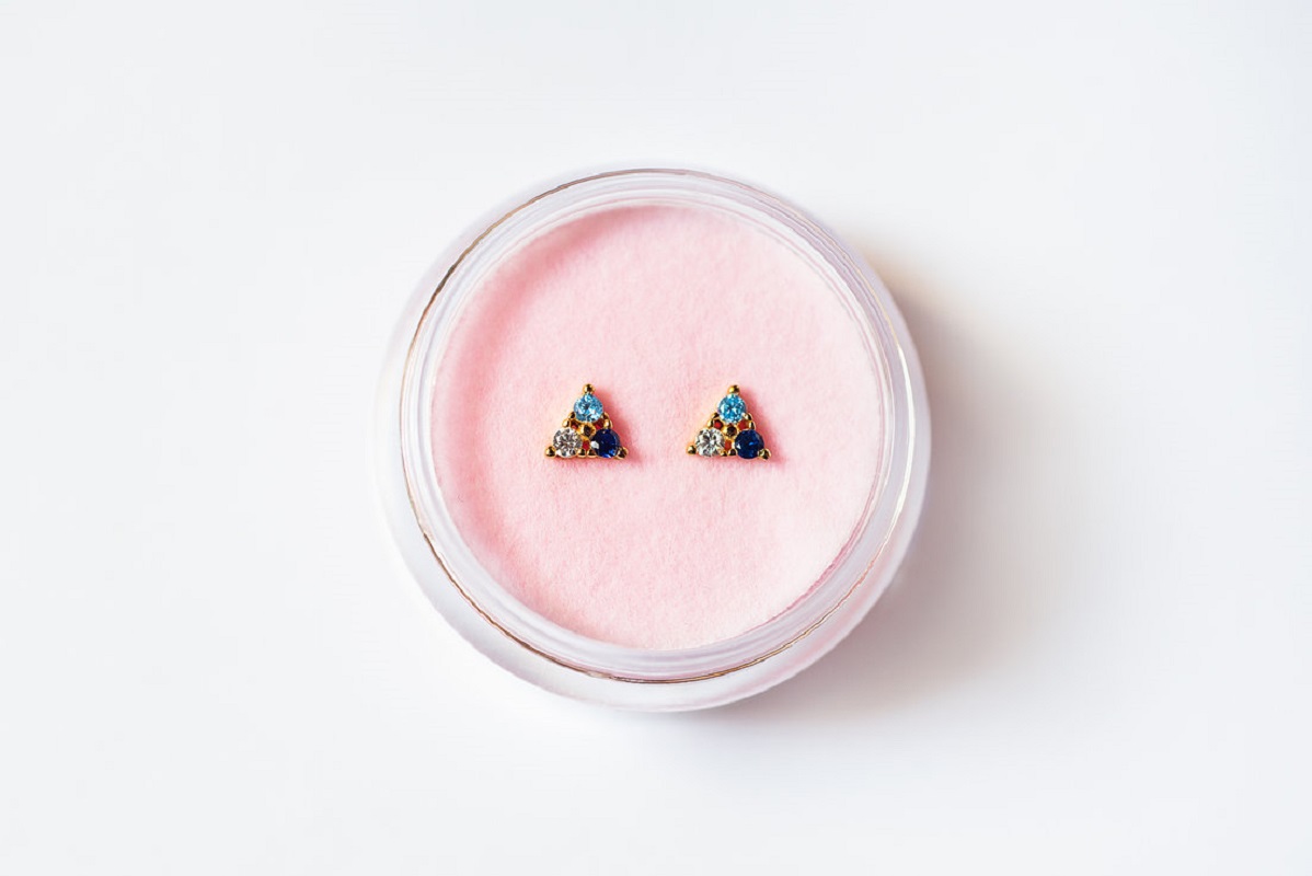 7a75ef_8b4ea4e9456a49d4839a1c195940d78f_mv2 Triangel-Ohrringe, goldfarben, blaue und weiße Steine, runde Form, rosa Hintergrund.