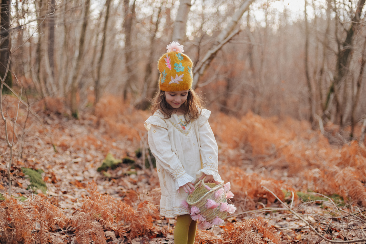 SKOVIA_CREAM_1 Kind mit beiger Tunika, orangefarbene Strickmütze mit bunten Blumenmustern, geflochtener Korb mit rosa Quasten, gelbe Strumpfhose.
