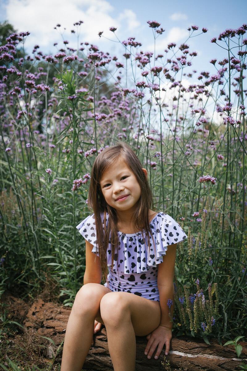 00008856568A472F_L Mädchenkleid; weiß mit schwarzen Herzmustern; Rüschenkragen; Sitzt inmitten blühender lilafarbener Blumen; natürliche Umgebung.