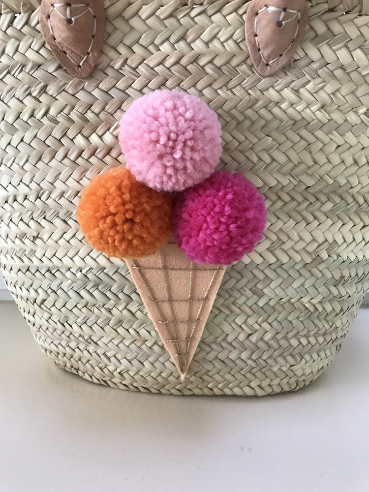 IMG_2582 Geflochtener Korb mit zwei braunen Lederschlaufen, dekoriert mit Eiscreme-Applikation aus drei bunten Pompons in Rosa, Orange und Pink als Kugeln, Eiswaffel aus beigem Stoff.