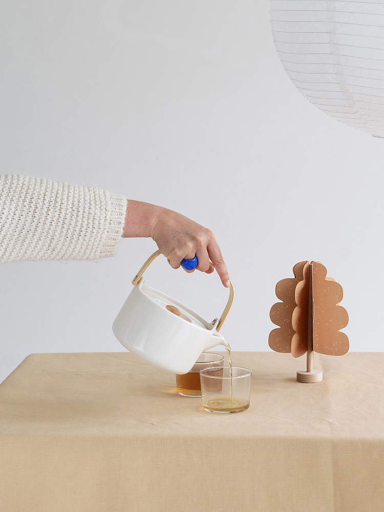 oak-tree-light-brown Weiße Teekanne, blauer Deckelgriff, Holzhenkel, zwei Glasbecher mit Tee, Tisch mit beigefarbener Tischdecke.