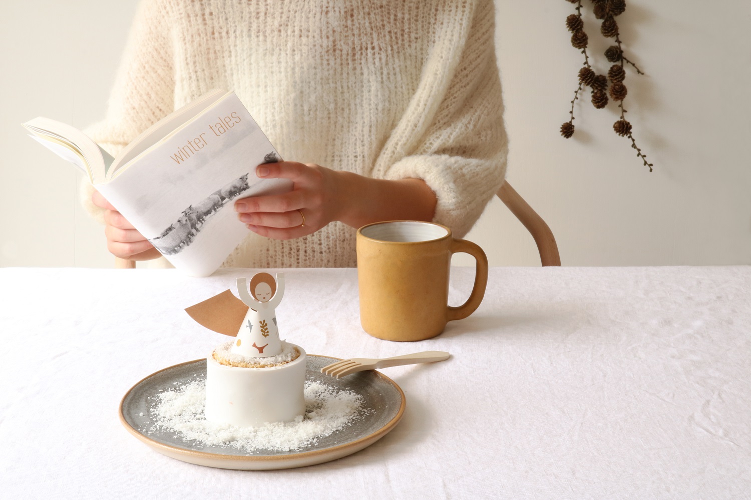 fridur-new-16 Kuchen mit Engel-Aufdruck, weiße Glasur, grauer Teller, beige Tasse, Person liest Buch „Winter Tales“, weiße Tischdecke.