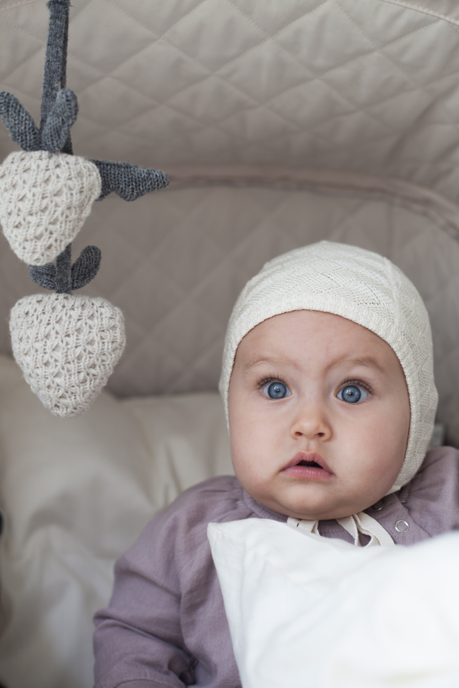 52 Bedienungsteile; Baby mit weißer Strickmütze; violetter Pullover; weiße Decke; Spielzeug-Mobile mit gestrickten weißen Beeren; graue Blätter; gesteppter Hintergrund.