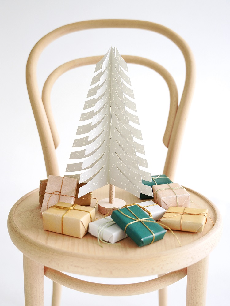 fir-tree-light-grey-1 Papier-Weihnachtsbaum, weiß mit Muster, auf Stuhl, umringt von kleinen, bunt verpackten Geschenken.