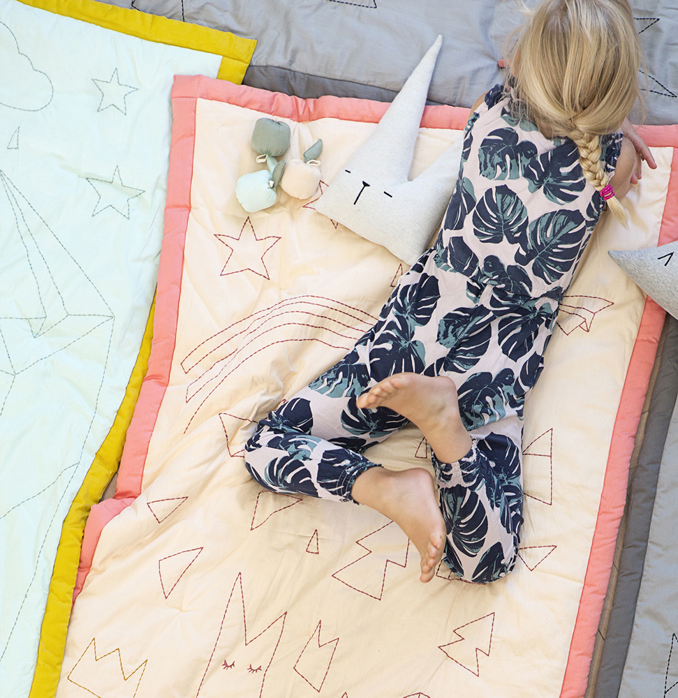 ON-A-JOURNEY-kids-1 Mädchen auf cremefarbener Decke mit rosa Rand, aufgestickte Sterne und Regenbogen, Pflanzenmuster-Pyjama, graues Kissen mit Gesicht, Spielzeug in Pastellfarben.