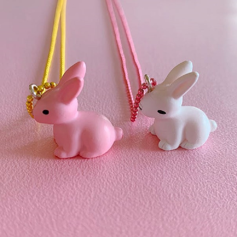 321423-47cd46bf116c65 Zwei Hasen-Anhänger; rosa Hase mit gelber Kette, weißer Hase mit pinker Kette; beide mit schwarzem Auge; rosa Hintergrund.