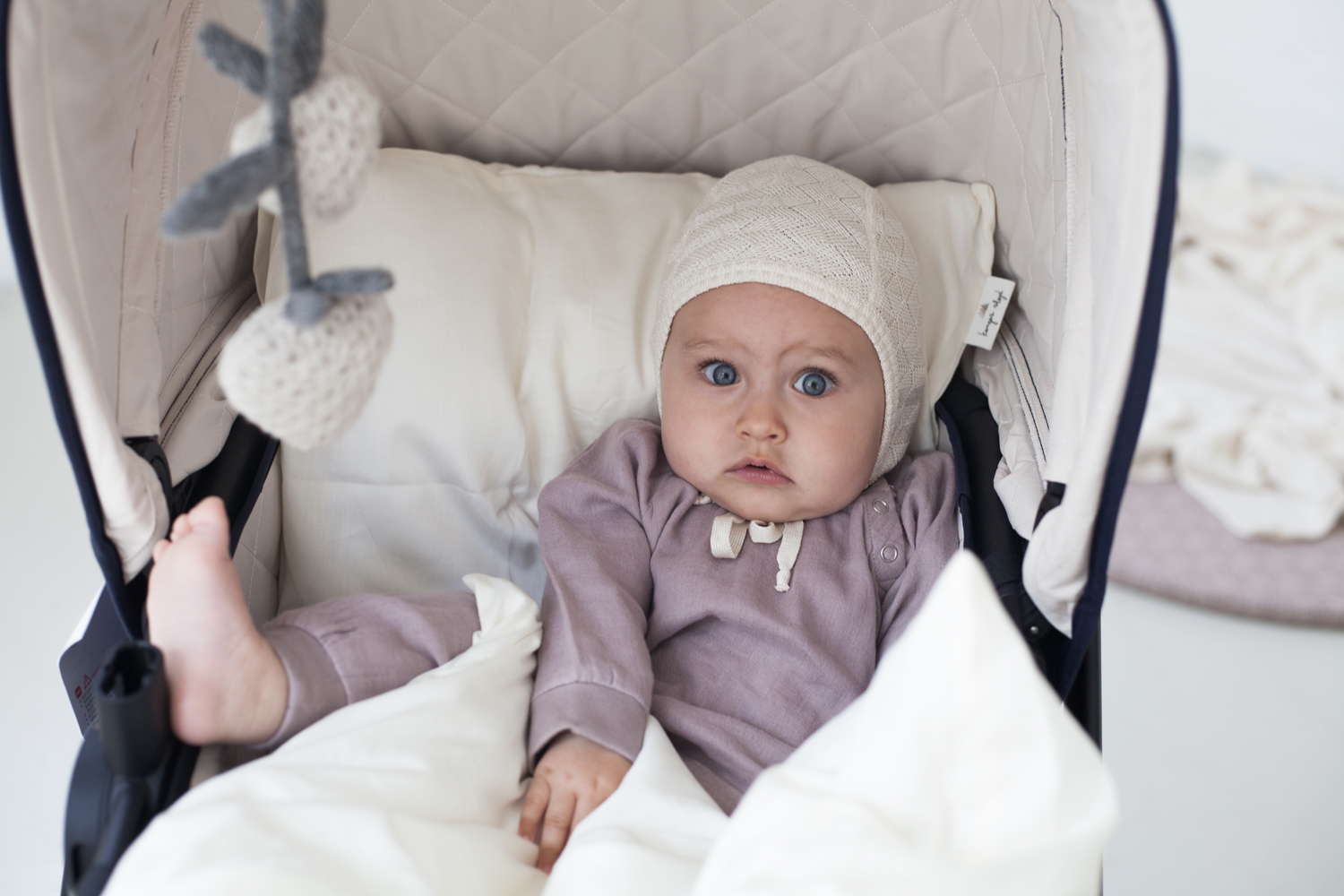 55 Kinderwagen, beige Innenpolsterung, Baby in lila Strampler, cremefarbene Strickmütze, weiße Decke, graue gehäkelte Anhänger.