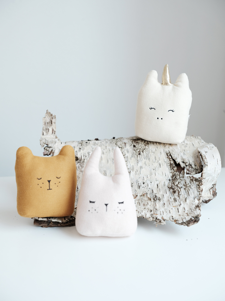 animal-music-mobiles-on-log Stofftiere, drei Stück; links ockerfarben, Bärngesicht; Mitte rosa, Hasengesicht; rechts cremeweiß, Einhorn, geschlossene Augen; alle auf einem Baumstamm platziert.