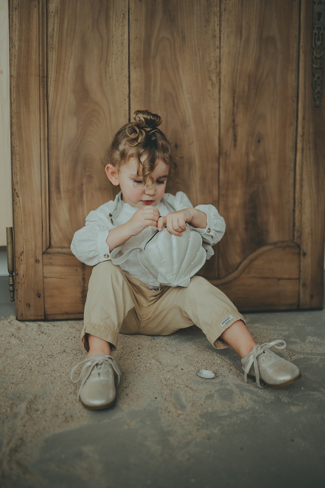 ANCK_donsje161-2 Kind mit weißer Bluse, beigefarbener Hose, silbernen Schuhen, hält eine kleine weiße Tasche. Hintergrund aus Holz.