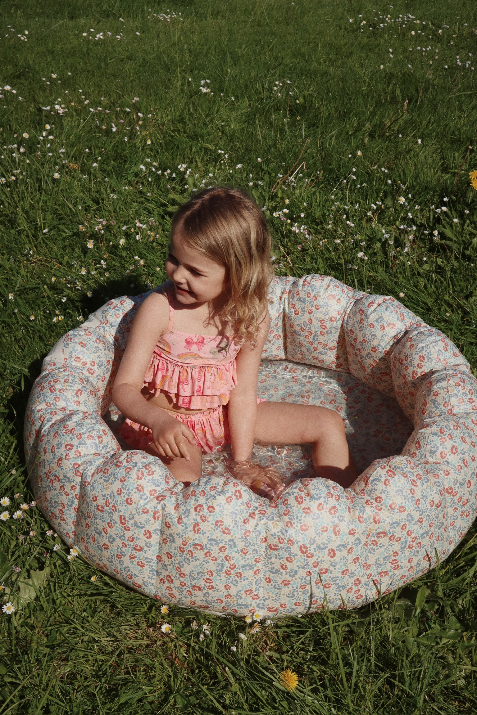 Aufblasbares Planschbecken, Blumendesign, bunt; Kind sitzt im Wasser; rosa Badeanzug mit Volants und Schleifenaufdruck.