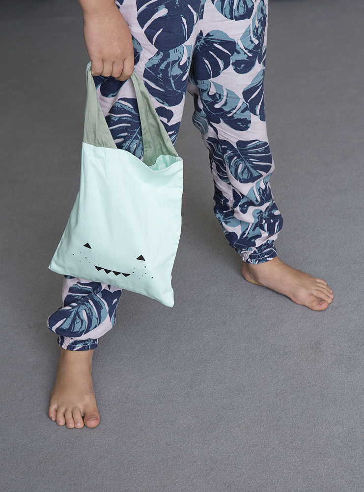 LUNCHBAG-ADVENTURER-kid-1 Kleine Stofftasche, hellgrün, Aufdruck Gesicht mit Dreiecken; getragen von Kind in Pyjama mit blau-grünem Blattmuster.