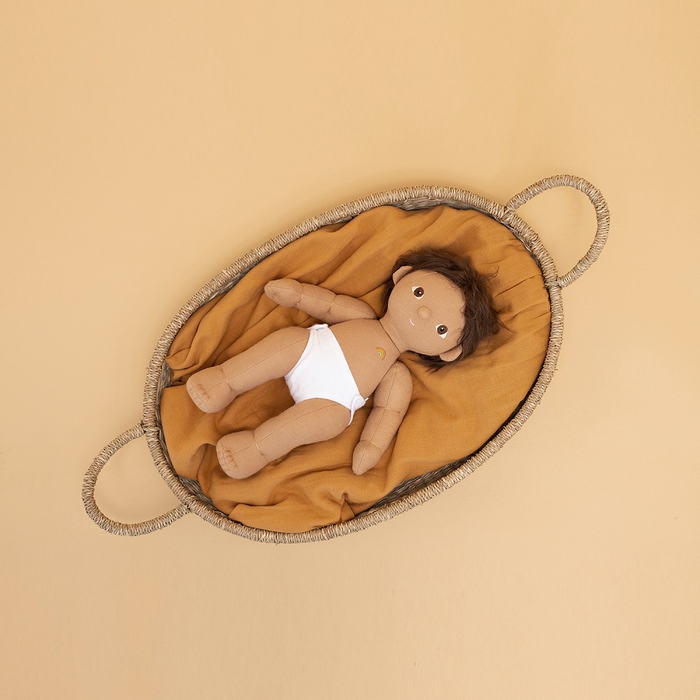 OE-Doll-Nyla Puppe im Körbchen, braune Haare, braune Haut, weiße Windel, kleiner Regenbogen-Aufdruck auf Bauch, oranges Innenfutter im geflochtenen ovalen Korb mit Griffen.