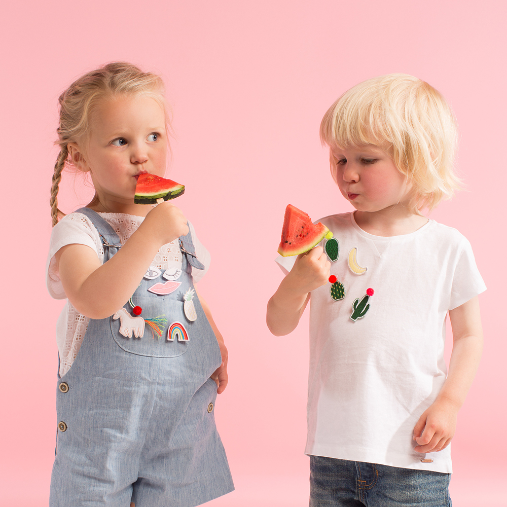 50-0084c Kleidung für Kinder, T-Shirt weiß mit Kaktus- und Fruchtapplikationen, Latzhose hellblau mit bunten Applikationen, beide Kinder mit Melonenscheiben.