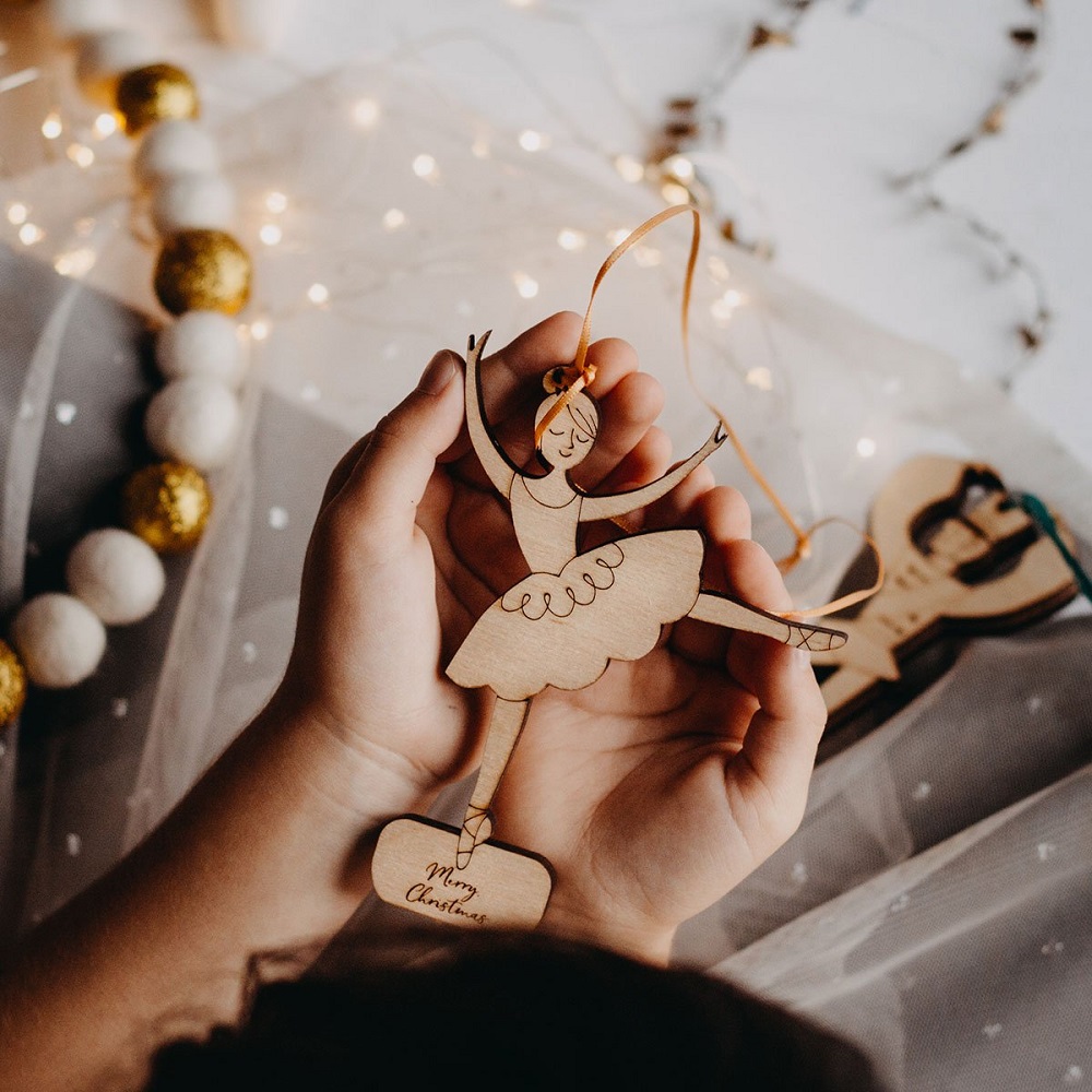 dancingballerina_1024x1024-2x Holzanhänger, Ballerina-Design, naturbelassen, Schleifenband, Schriftzug "Merry Christmas" am unteren Ende, in Kinderhand gehalten.