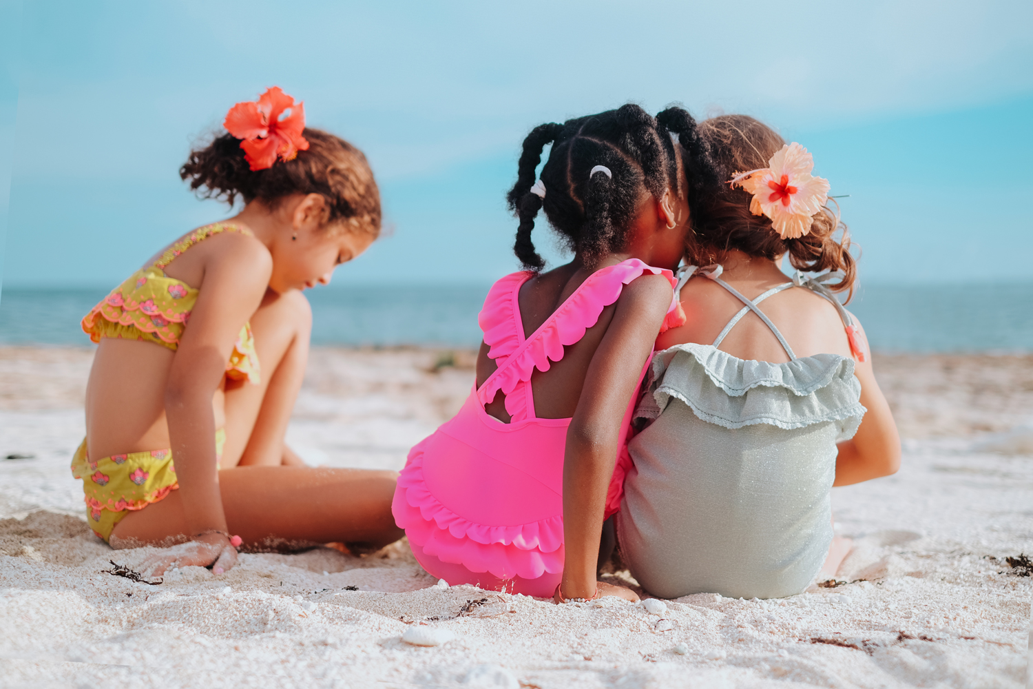 kidsbathingsuitcreolealmond8 Mädchen-Badebekleidung, gelbes Bikini-Set mit rosa Rüschen, pinker Badeanzug mit Rüschen, hellblauer Badeanzug mit Rüschen, Blumen im Haar, Strandumgebung.