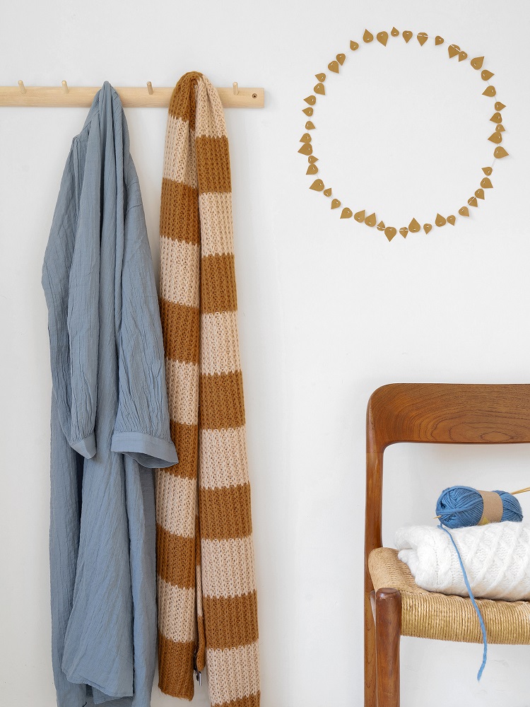 twig-leaves-ochre-2 Kleiderhaken mit blauem Stoff und gestreiftem Schal, braun-beige, darüber Wanddeko aus Holz; daneben Stuhl mit weißer Decke und blauem Wollknäuel, Stricknadeln.