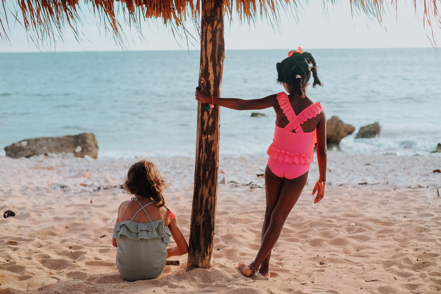 kidsbathingsuitcreolealmond10 Zwei Kinder am Strand; grauer und pinker Rüschen-Badeanzug; Sand, Meer im Hintergrund.