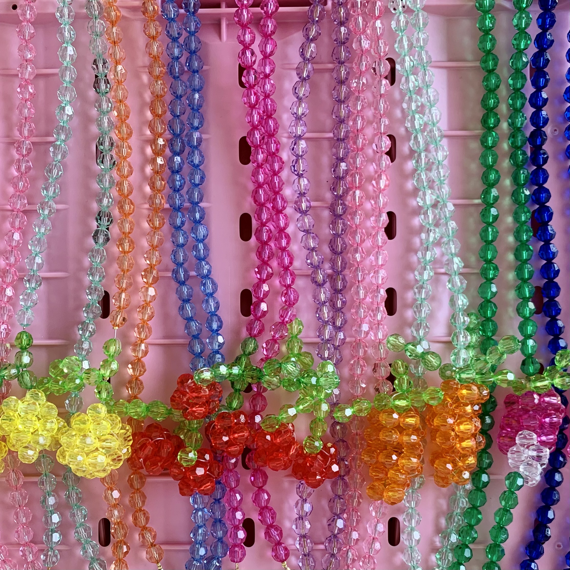 Bunte Perlenketten, auf rosa Hintergrund, viele Farben: pink, grün, blau, lila, gelb; einige Ketten mit Blumen- oder Fruchtanhängern; detaillierte, facettierte Perlenstruktur; transparente Optik.