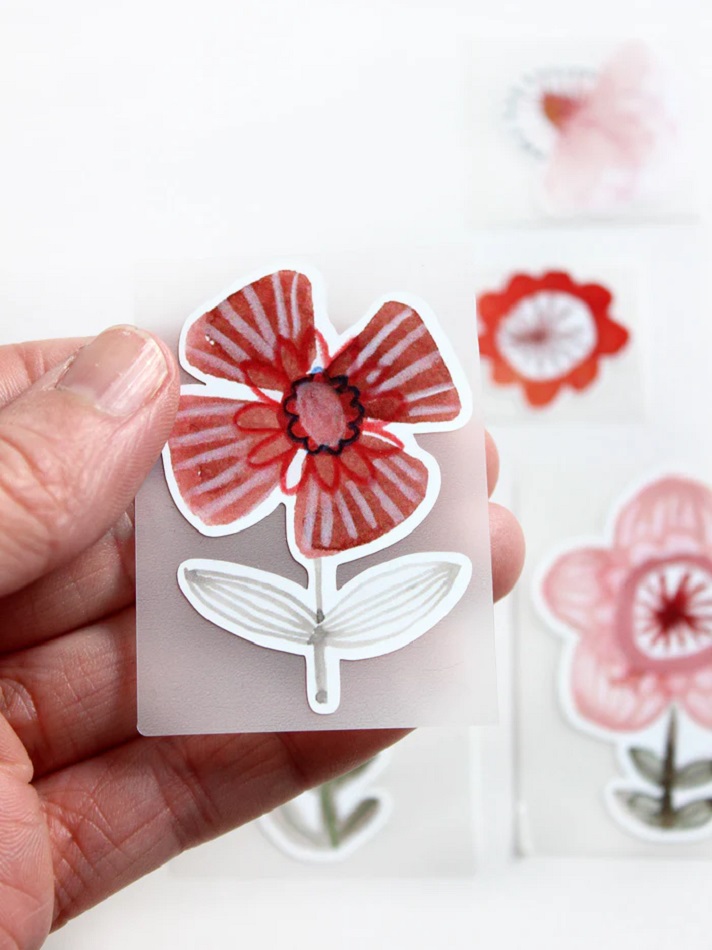 Buegelbilder_Set_Bluetenundblumen_halfbird3_700x Sticker, rote Blume, grüne Blätter, transparente Umrandung, Hand hält Sticker, unscharfe Hintergrundmotive.