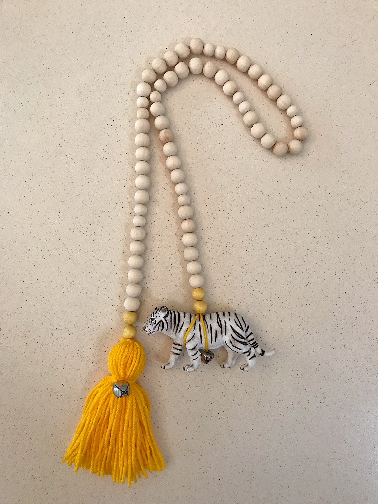F00284DE-0175-4ED1-96B6-83418A134F3C-002 Holzperlenkette, naturfarben, gelber Quastenanhänger, weißer Tigeranhänger mit schwarzen Streifen, kleiner silberner Glöckchencharme.