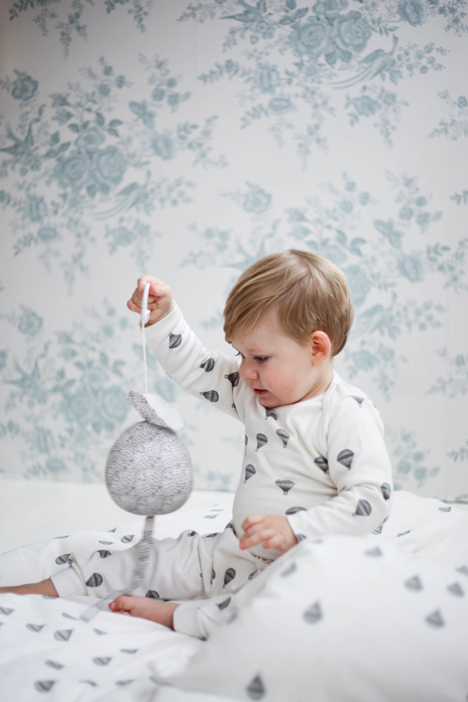 500007-MARSHMALLOW-MUSIC-MOBILE Baby-Pyjama, weiß, graue Heißluftballon-Aufdrucke, gehäkeltes Vogel-Mobile in Grau, hellblaue und graue florale Tapete im Hintergrund.