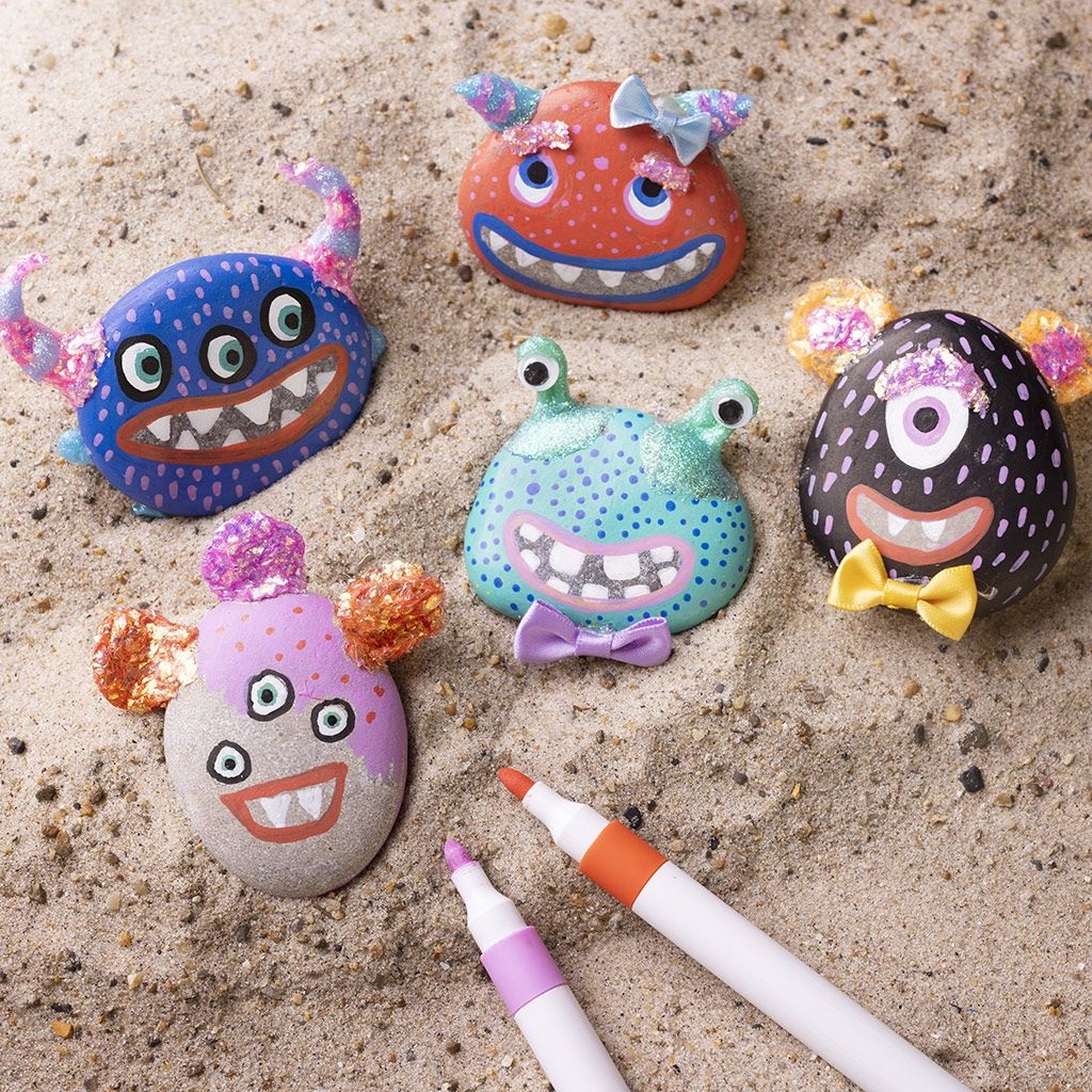 Bemalte Steine, Monsterdesigns, leuchtende Farben, Glitzerakzente, Schleifen und Ohren, bunte Tupfenmuster, zwei farbige Marker (lila, orange) im Vordergrund, Sandhintergrund.