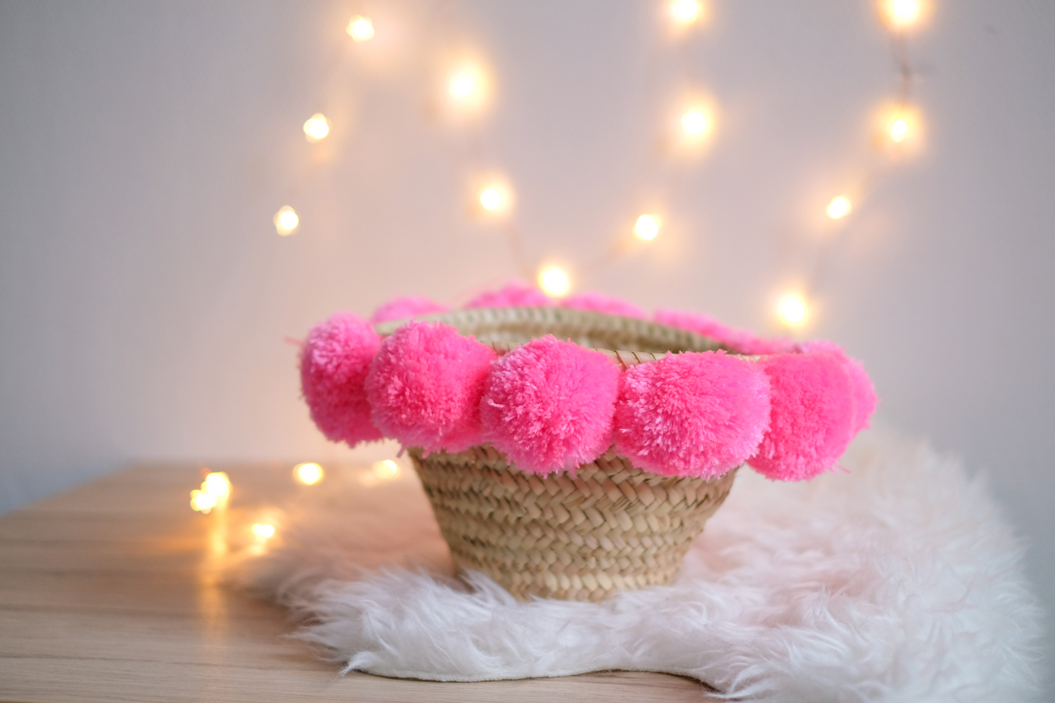 KYRLA_FLUOPINK_1 Geflochtener Korb, beige; dekoriert mit pinken Pompoms; rund; auf weißem Fell.