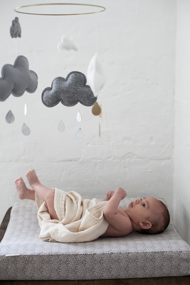 10159ce54a148479 Baby auf beige gepolsterter Wickelauflage, Mobiles aus grauen und weißen Stoffwolken mit Regenform-Anhängern, neutrale Wände.