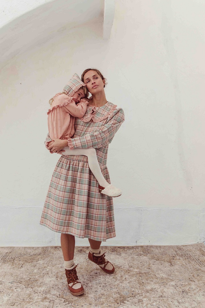 LIAW21_40_W_check_mama-bebe Frau in karierter Kleid, Rüschenkragen, knöchellang; Kind in rosa Kleid mit Kapuze, weißer Strumpfhose; beide barfuß; rustikaler Hintergrund.