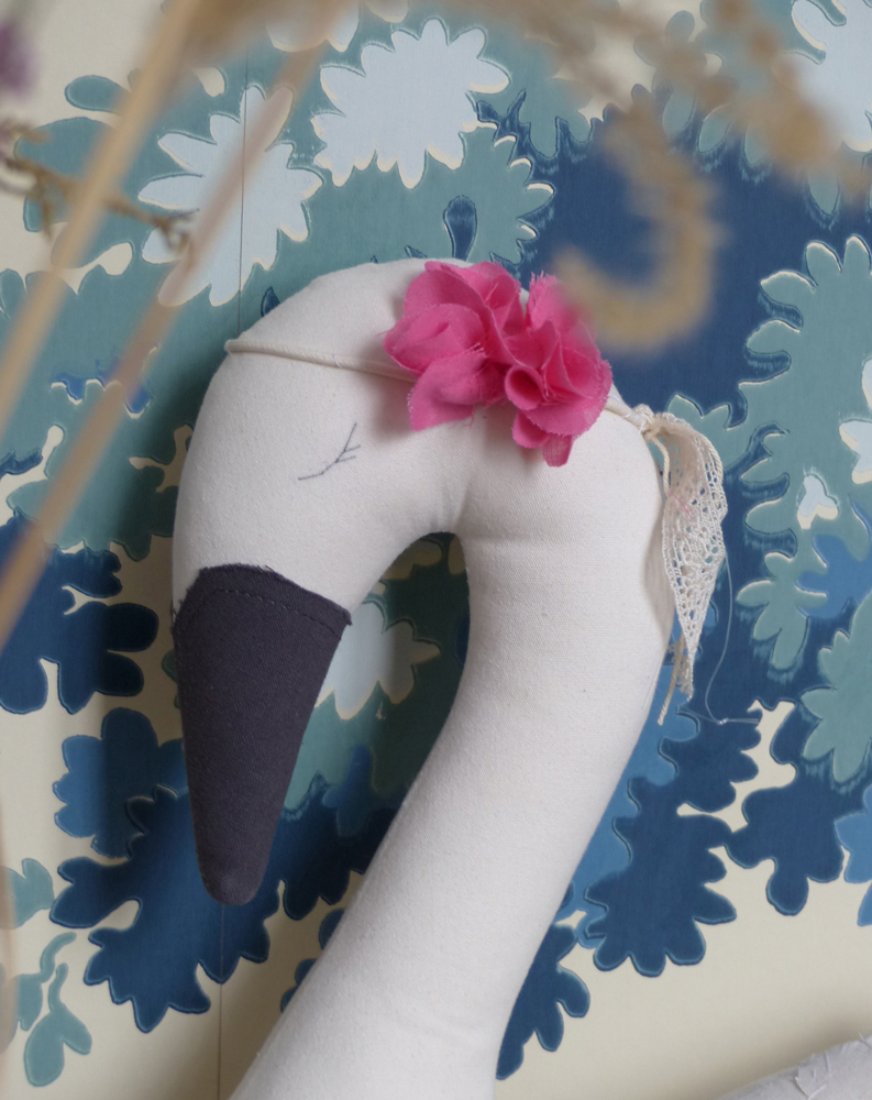 Scala-Swan-le-cygne-1 Stofftier Schwan, weiß, schwarzer Schnabel, rosa Blumenkranz, Spitze, blau-weißer Hintergrund, florales Muster.