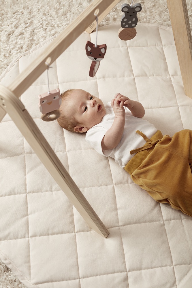 1000483-Play-mat-1000365-Baby-gym-1000416-Figures-EWM_1 Babyspielbogen aus Holz, helles Holz, hängende Tierfiguren in Grau und Braun, Baby liegt auf cremefarbener, gesteppter Spielmatte, Baby in weißem Shirt und brauner Hose.