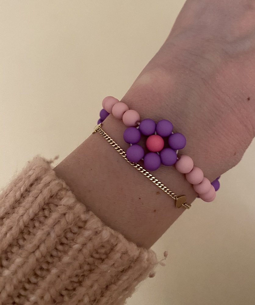 IMG_5136 Armband-Set, eine Kette aus Gold mit Herz, Perlenarmband in Lila und Pink mit Blumenmuster, getragen auf Handgelenk mit beigem Strickpullover.