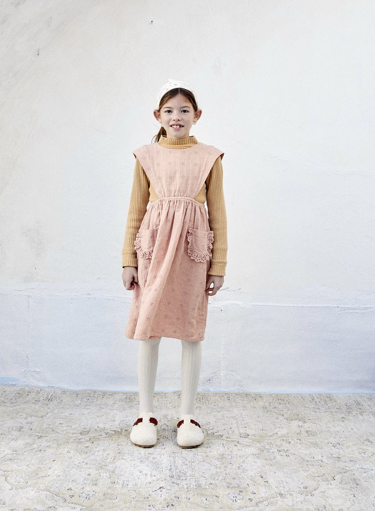 Mädchenkleid, rosa mit floralen Mustern, Rüschen an Taschen, darunter Rollkragenpullover beige, weiße Strumpfhose, flauschige Hausschuhe cremefarben mit braunen Details, Haarband weiß.