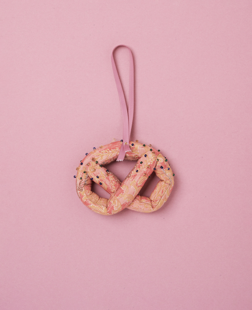 Pretzel-SILK-Ornament-Pinkie-Pie Stoffbrezel, rosa Marmor-Muster, bunte Stecknadeln, rosa Band, Hintergrund rosa.