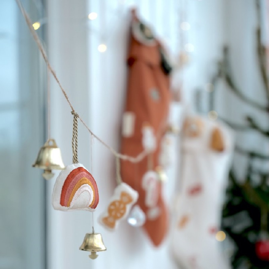 FBL-Ornaments-Hanging-Rainbow-Candy-1 Glockengirlande, Stoffanhänger, Regenbogenmotiv, orange-weiß, unscharfe Weihnachtsstrümpfe im Hintergrund.
