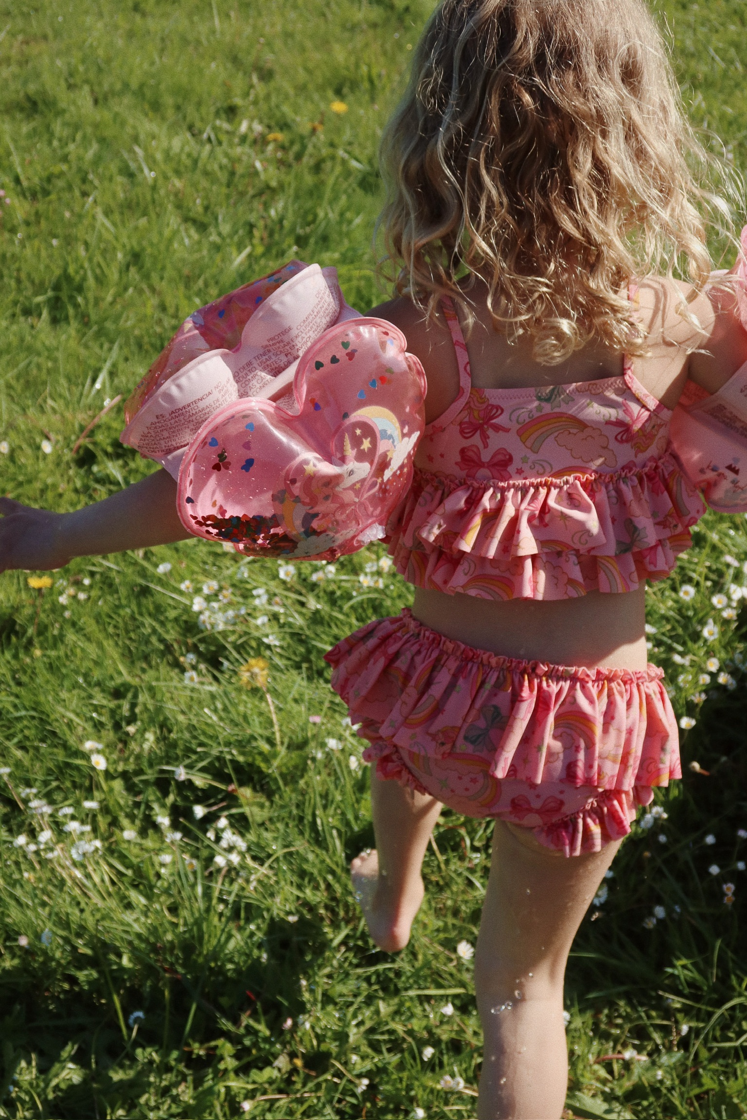 Kinder-Badeanzug, rosa, Rüschen, Einhorn- und Regenbogenmuster, dazugehörige rosa Schwimmflügel mit bunten Glitzerherzen.