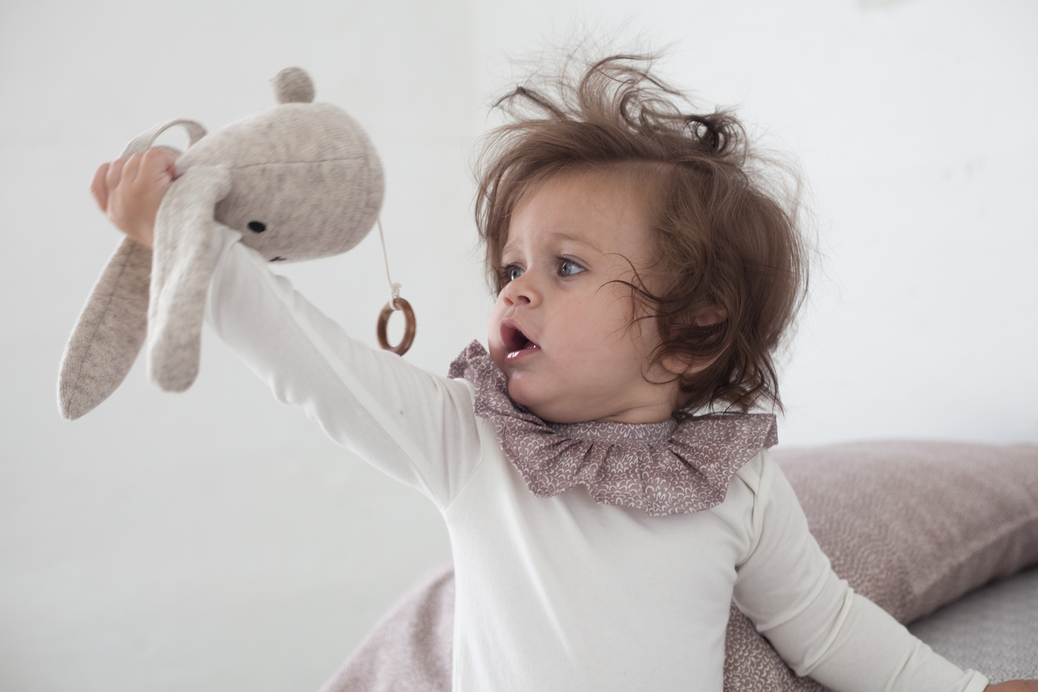 0558c1573a08ef1 Stofftier Hase, beige, mit Holzring; Baby in weißem Langarmshirt mit gemustertem Kragen.