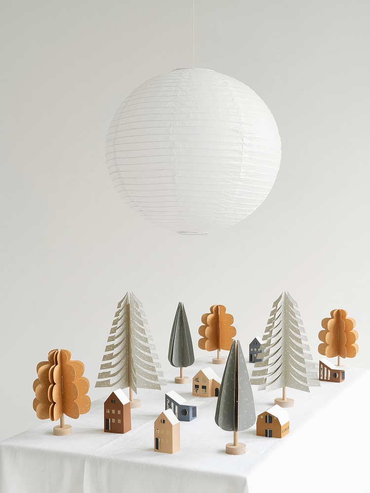 oak-tree-light-brown-1 Papierlampion, rund, weiß; Holzfiguren: Tannenbäume grün, braun, weiß; Häuser beige, braun, blau mit weißen Dächern; Auf Tisch vor weißem Hintergrund.