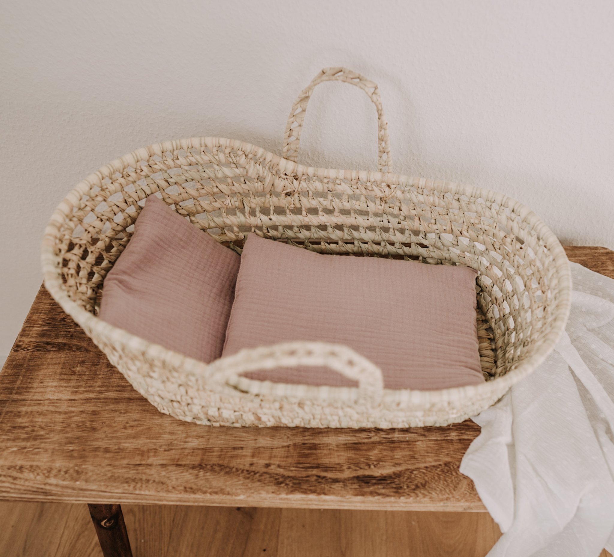 image_9d21df70-cb44-422d-bbe6-f73d10909193_1024x1024-2x Geflochtener Korb, beige, zwei Griffe, rechteckige Form; innen zwei rosa Kissen; auf Holzoberfläche.