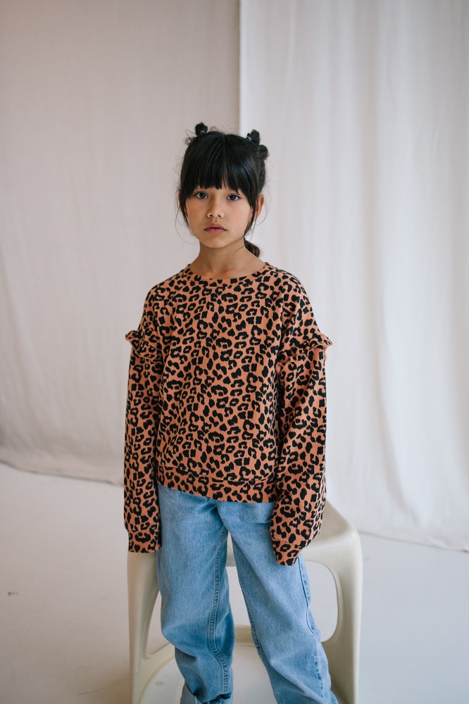2-25 Kinderpullover, Leopardenmuster, braun-schwarz, lange Ärmel, Rüschen an den Schultern; blaue Jeans; weißer Stuhl.