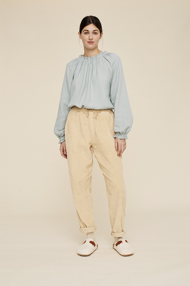 liaw22_076_W_Nola-Blouse-Woman_aqua-grey-1 Damenoutfit, hellblaue Bluse mit Rüschenkragen, beiger Cordhose, weiße Hausschuhe mit braunem Detail.