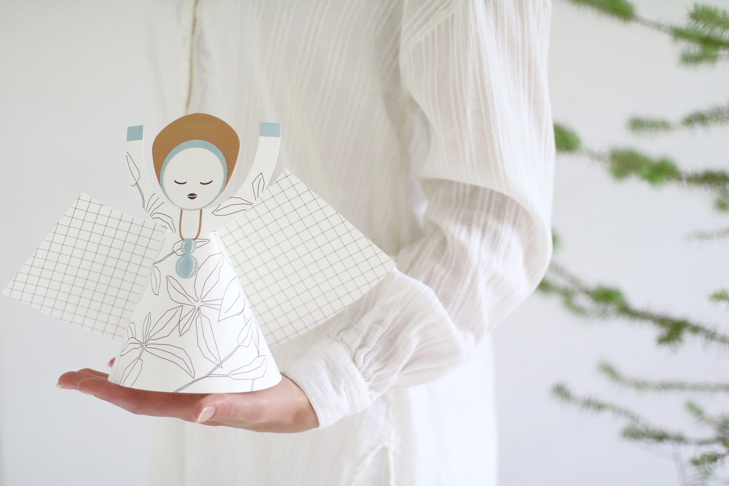 XL-angel-hand Papierengel, weiß mit Pflanzenmuster, rechteckige Flügel mit Karomuster, rundes Gesicht, braune Kapuze, gehalten in Hand, dezentes Design.