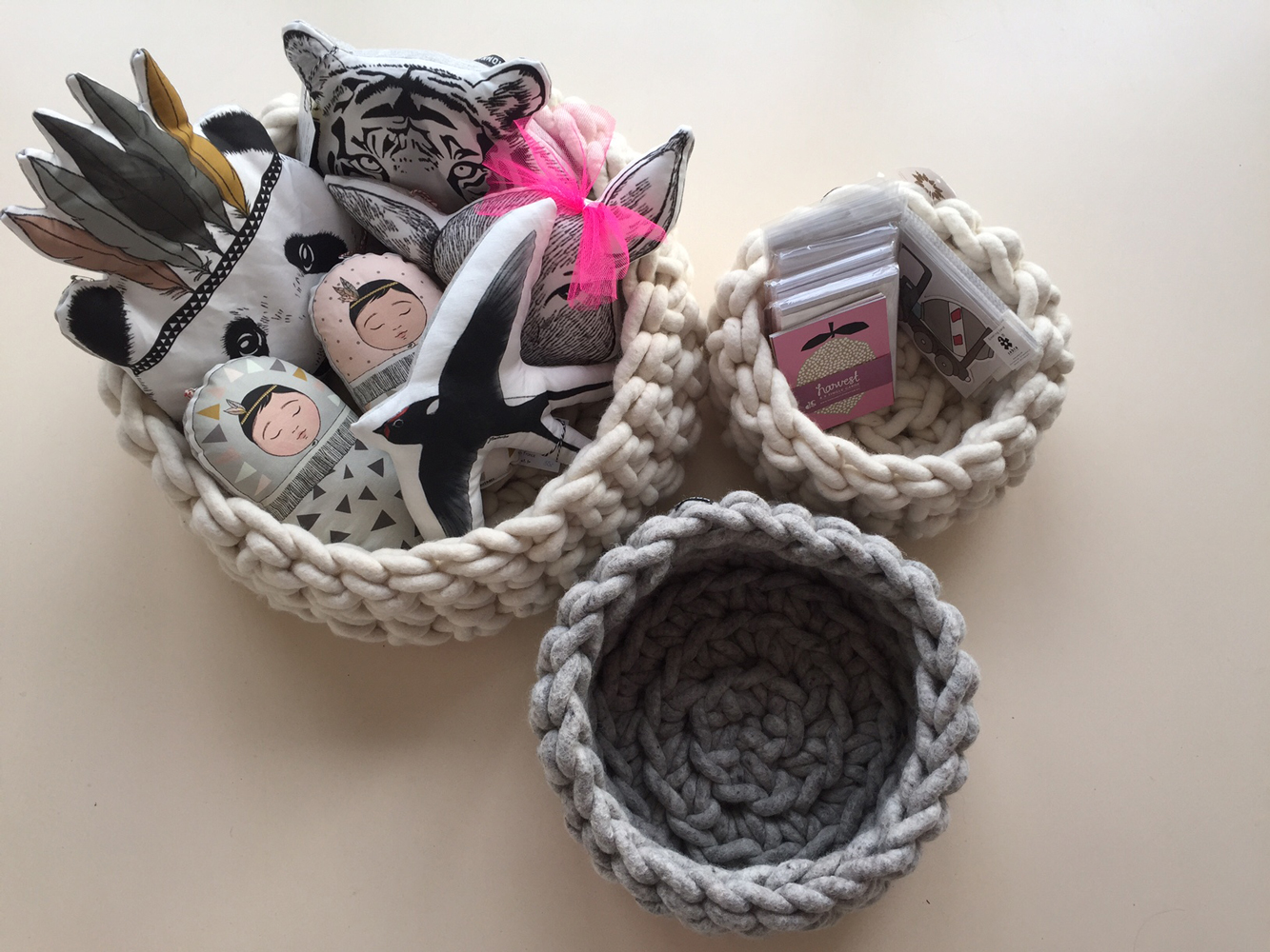 IMG_8257 Gehäkelte Körbe, creme und grau; gefüllt mit bunt bedruckten Stofftieren, Federn, Tigermuster, Gesichter; kleine Kartenstapel, teils rosa Verpackungen.