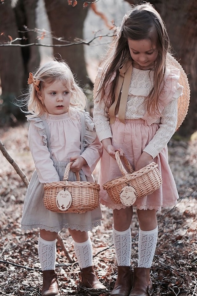 easterisabel14_1024x1024-2x Zwei geflochtene Kinderkörbe mit runden Holzschild-Anhängern, getragen von Mädchen in Spitzenkleidung; Körbe naturfarben, Anhänger mit Aufdruck.
