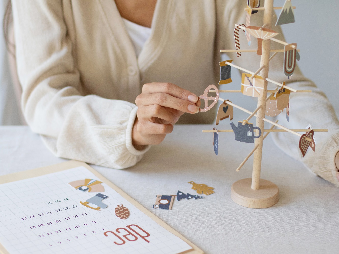 advent-tree-26 Holzständer mit festlichen Papieranhängern; Motive: Brezel, Herz, Teekanne, Geschenk; darunter Kalenderblatt; Person in heller Kleidung arrangiert Dekoration.