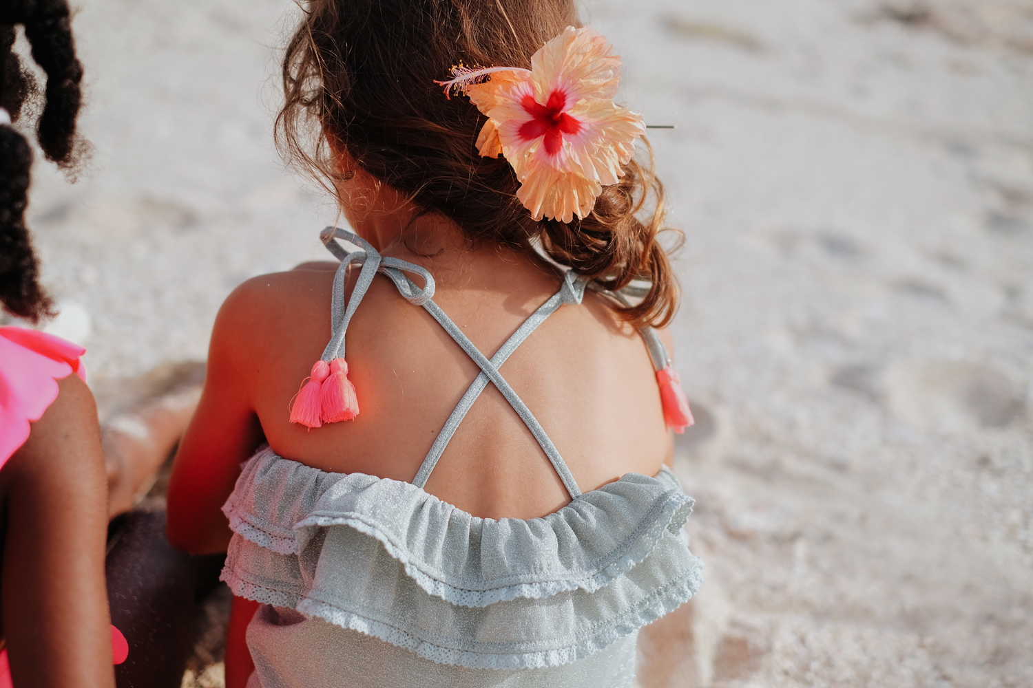 kidsbathingsuitcreolealmond2 Kind in Sommerkleid mit Rüschen, blassblaue Farbe, gekreuzte Träger, pinke Quasten, große Blume im Haar.