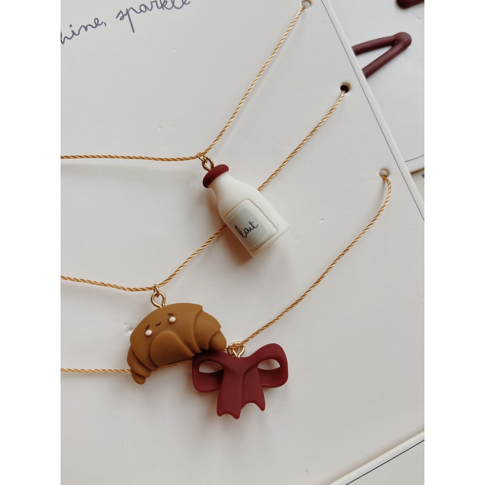 1024x1024-7da36980c134a907d6575c0c1883d6ce Drei Armbänder mit Anhängern: weiße Milchflasche mit "lait"-Aufschrift, braunes Croissant, dunkelrote Schleife; goldene Ketten.