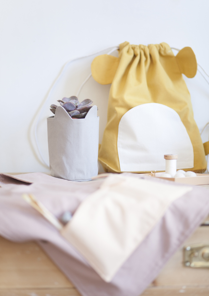 IMG_8564591acba60ba31 Stoffbeutel gelb mit Tierohren, cremefarbenes Rundmuster; grauer Topf mit Pflanze; Pastellfarbene Textilien im Vordergrund; Garnrollen.