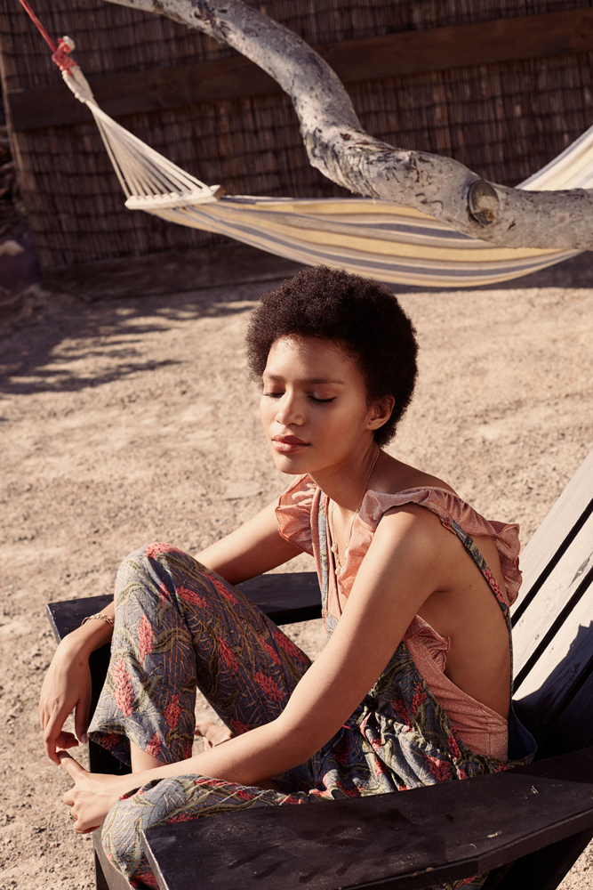 womenbathingsuitdorevarusty7 Frau in gemusterter Sommerhose und rosa Oberteil mit Rüschen; dunkle, kurze, lockige Haare; sitzt auf Holzstuhl im Freien.