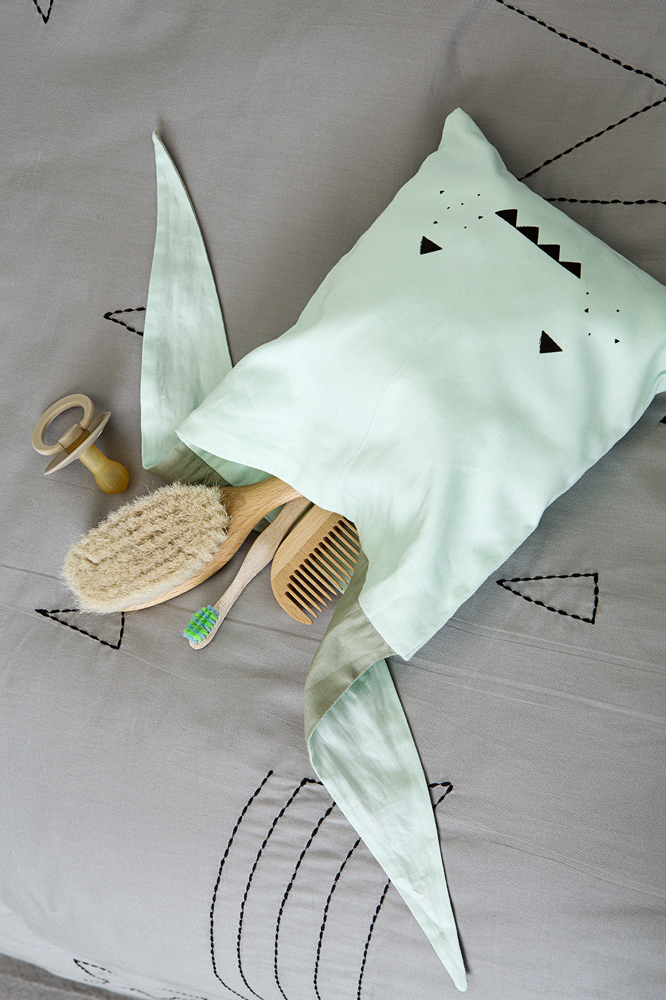 lunchbag-adventurer-2 Kissen mintgrün, Aufdruck schwarzes Gesicht mit Dreiecken; Holzbürste, Holzhaargriffkamm, Zahnbürste mit grünem Kopf; Schnuller mit Holzgriff; graue Unterlage mit geometrischen Mustern.